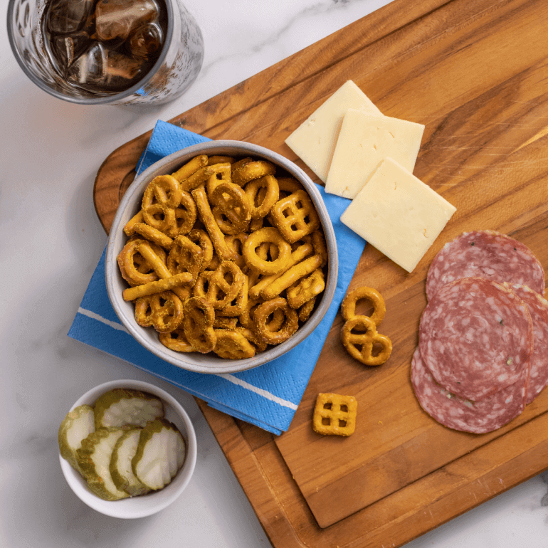 A bowl of pretzels on a cutting board with cheese, meat and pickles