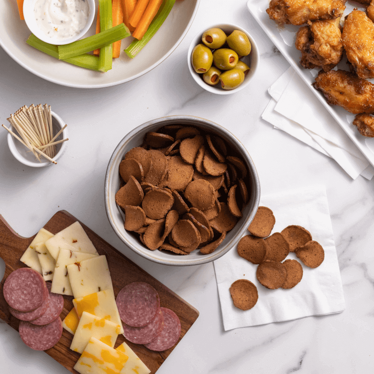 A bowl Rye Chip Gardetto's with a veggie plate, cheese plate, chicken wings and olives all on a table