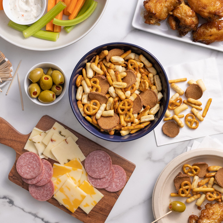 A bowl of Gardetto's snack mix in the middle, surrounded by a veggie plate, meat and cheese charcuterie board, chicken wings and olives on a table